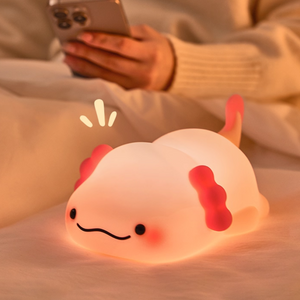 Cute axolotl-shaped night light on a soft surface with a person using a phone in the background.