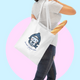 White tote bag with cute shark kitty illustration, blue text, carried by a young lady ideal for adding playful style to shopping. Tote bag