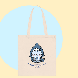 Cream tote bag featuring a kawaii kitty in a blue shark costume, playful slogan, and green trainers—perfect for everyday use. Tote bag