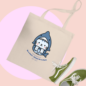 Cream tote bag featuring a kawaii kitty in a blue shark costume, playful slogan, and green trainers—perfect for everyday use. Tote bag